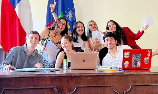 Grupo de siete estudiantes, posando con una sonrisa, con dos computadores al frente y las banderas de Chile y la Universidad de Chile, detrás