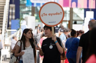 Monitor Uchile, con cartel que dice "¿Te ayudo?", conversando con una postulante, en la Feria del Postulante 2019