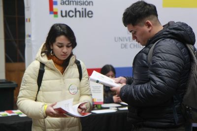 Una y un estudiante, ambos con parka, leen documentos informativos. Atrás, en un pendón, se ve el logo de Admisión Uchile.