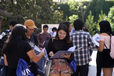 Grupo de estudiantes de pie en un patio. De frente se ve a una estudiante sonriendo, mirando una carpeta. Foto corresponde al archivo de una actividad de Inducción a la Vida Universitaria