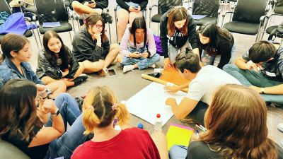 Estudiantes jóvenes, en círculo en el suelo, compartiendo y trabajando en un papelógrafo.