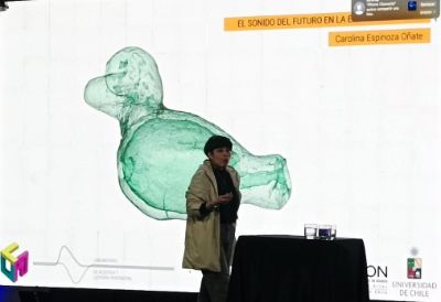 La profesora Carolina Espinoza, exponiendo, y en el fondo proyectado un modelo tridimensional de un silvato latinoamericano
