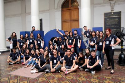Grupo numeroso de estudiantes monitoras y monitores, con polera de la Uchile, posando al frente de letras volumétricas UCH (180 cm de alto), al interior de Casa Central de la U. de Chile