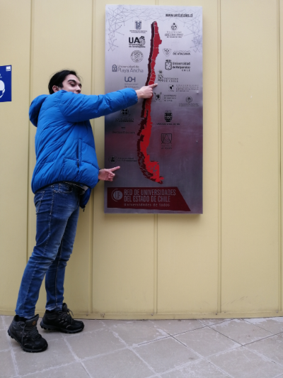 Nicolás Silva posa frente a placa de la red de universidades del Estado de Chile, donde aparece el mapa del país, con cada una de ellas. Él apunta a la U. de Chile y la U. de Magallanes.