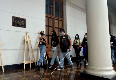 Estudiantes caminan por pasillo de la casa central, con mascarillas y uno de ellos, con la guía de una de sus compañeras y su bastón para personas con ceguera.