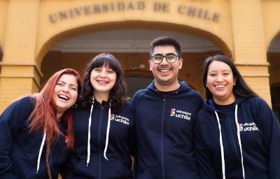Estudiantes con polerones de admisión Uchile, sonriendo frente a la fachada de FAU donde se alcanza a leer detrás Universidad de Chile 